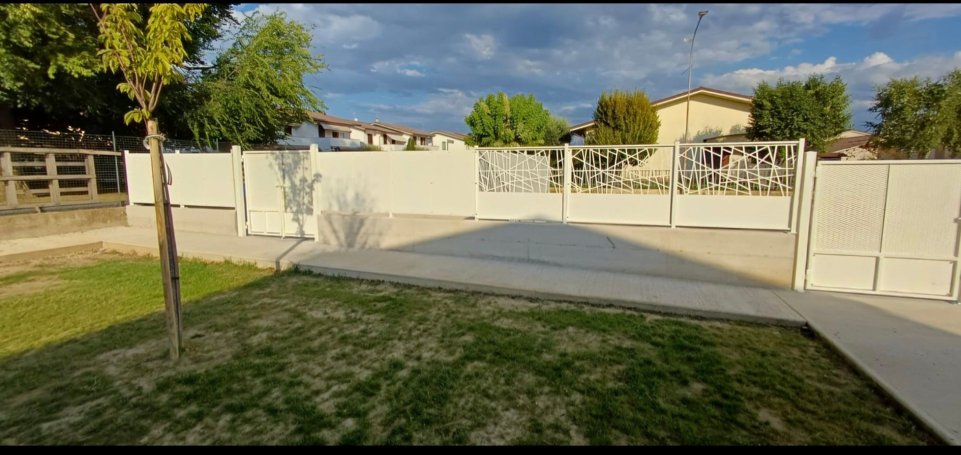 Ingresso con cancello bianco, prato verde e alberi, case sullo sfondo e cielo nuvoloso.