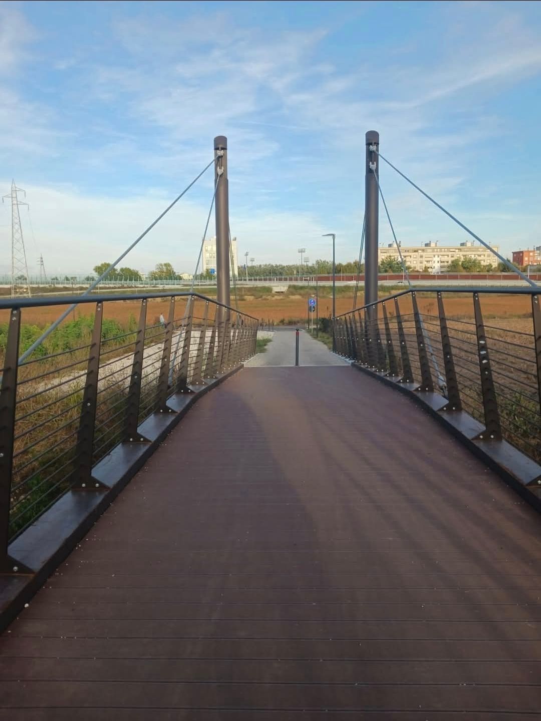 Un ponte pedonale in legno sopra il letto asciutto di un fiume, che collega due zone. Giornata di sole, cielo azzurro.