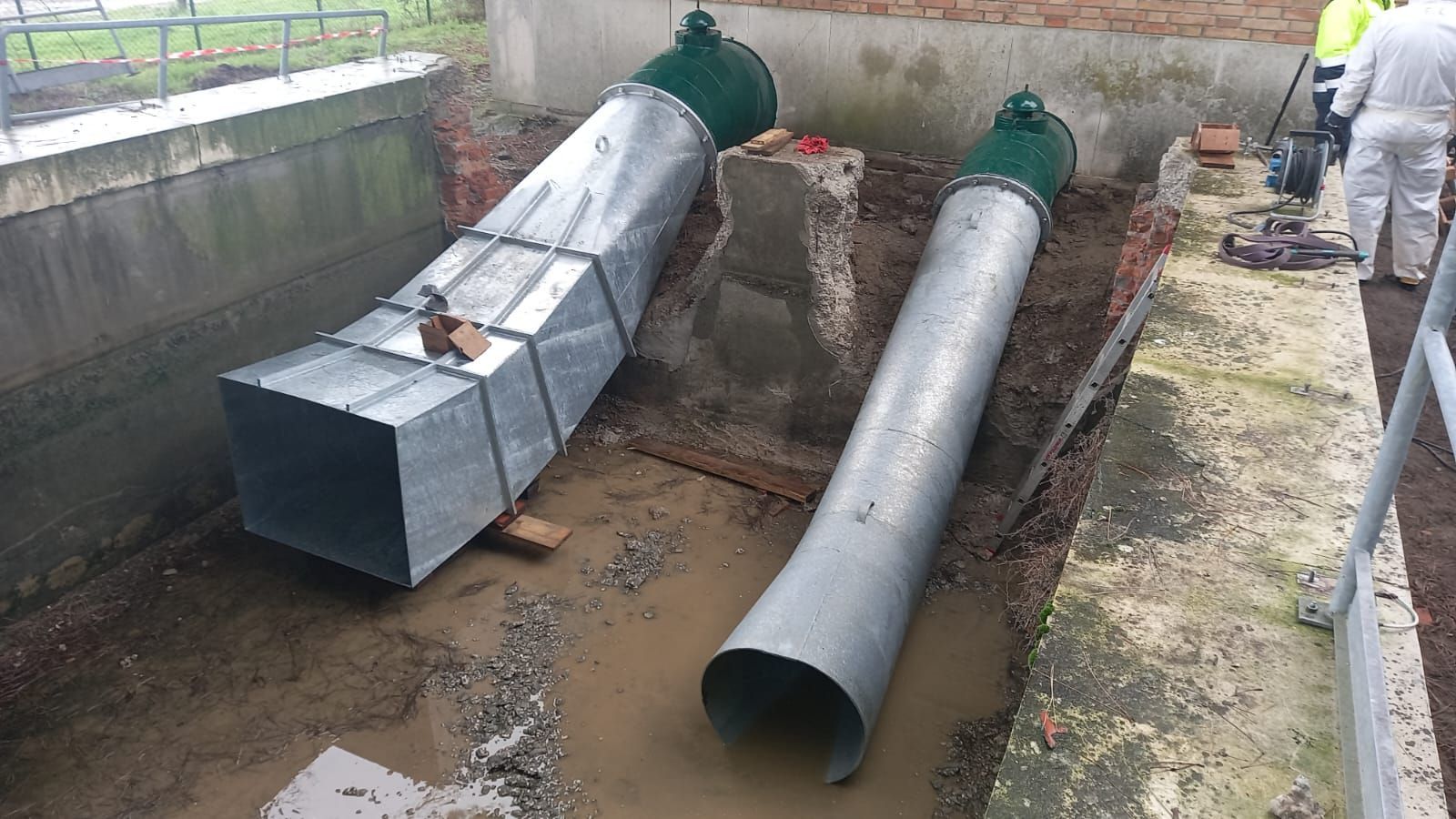 Due grandi tubi metallici e un operaio vicino a un muro di mattoni, in una zona fangosa.