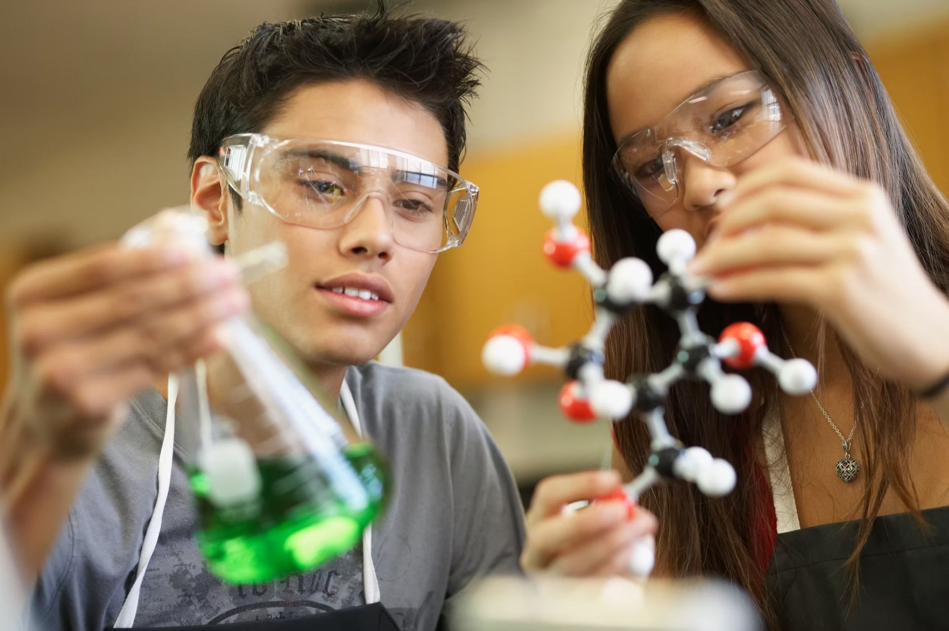 Two students in lab coats and safety glasses examine lab equipment and a molecular model.