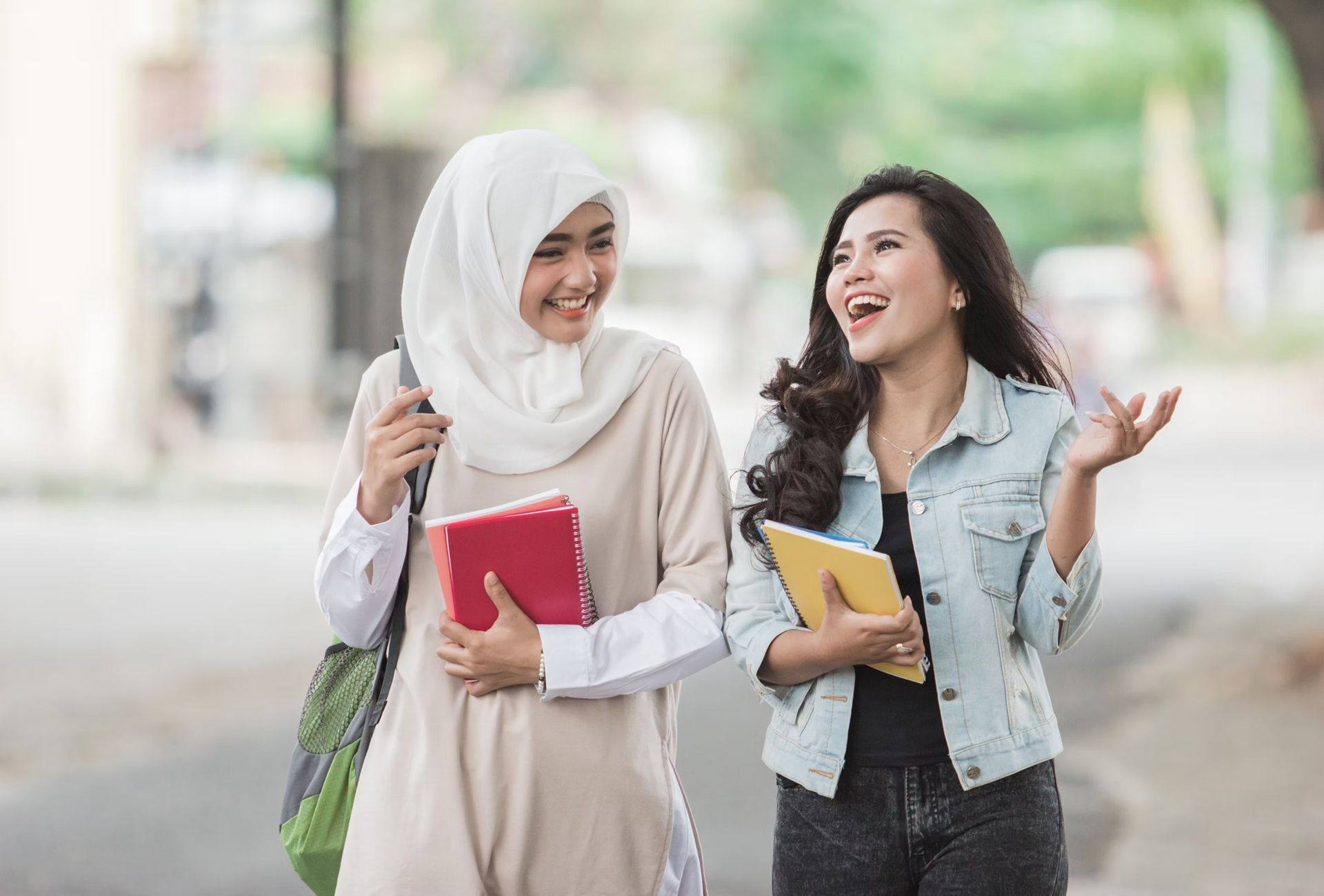 Two women walking and laughing on a street, one in a hijab and the other in a jean jacket, holding books.