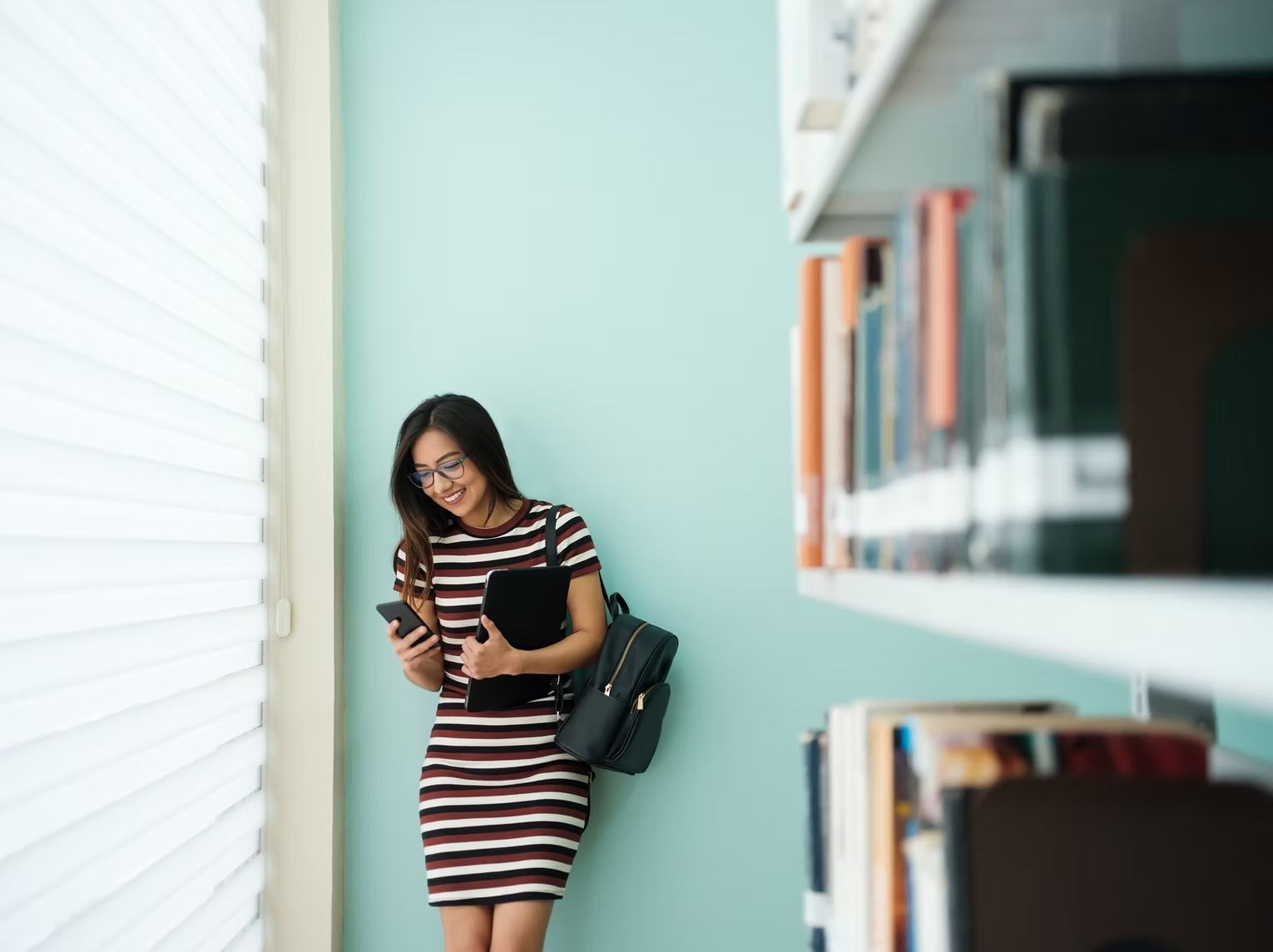 Woman with glasses, smiling, looks at phone in library. She wears a striped dress and backpack near bookshelves.