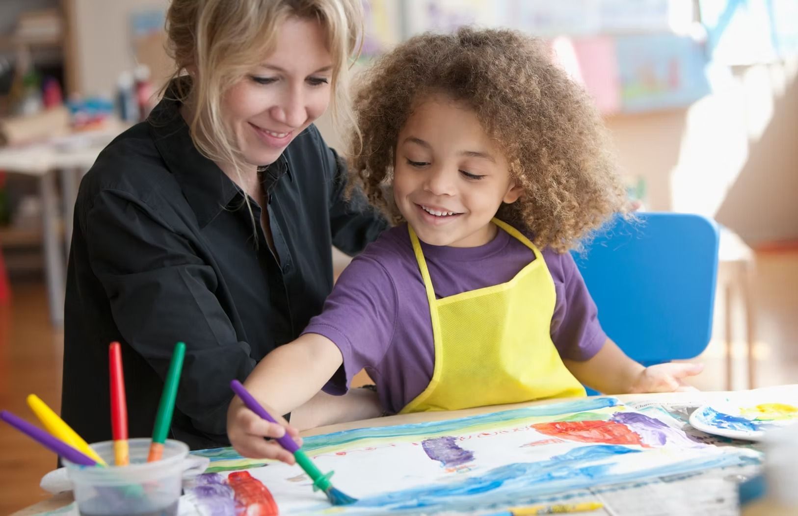 Woman assists child painting; both smiling at a table with paints and brushes in an art classroom.