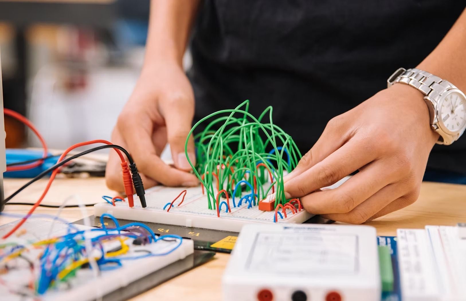 Person working on a circuit board with green and red wires, hands focused, watch on wrist.