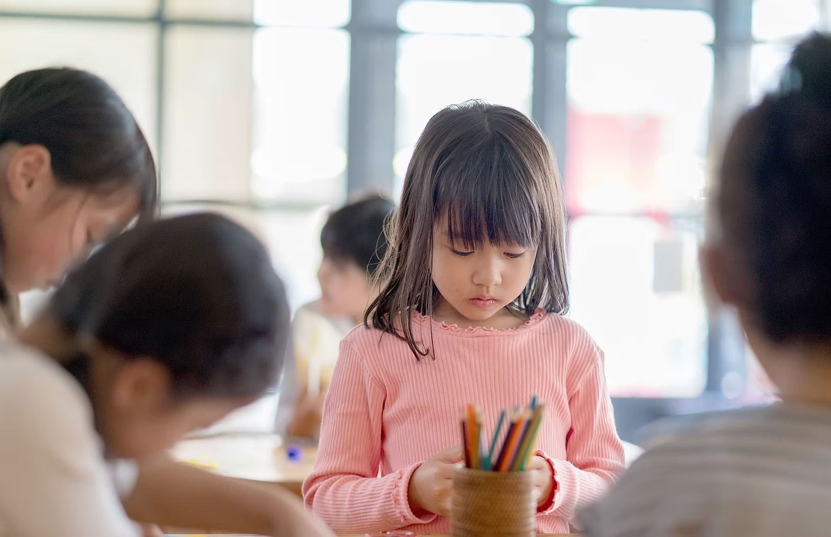 Girl in pink shirt holding crayons, looking down in a classroom setting with other children.