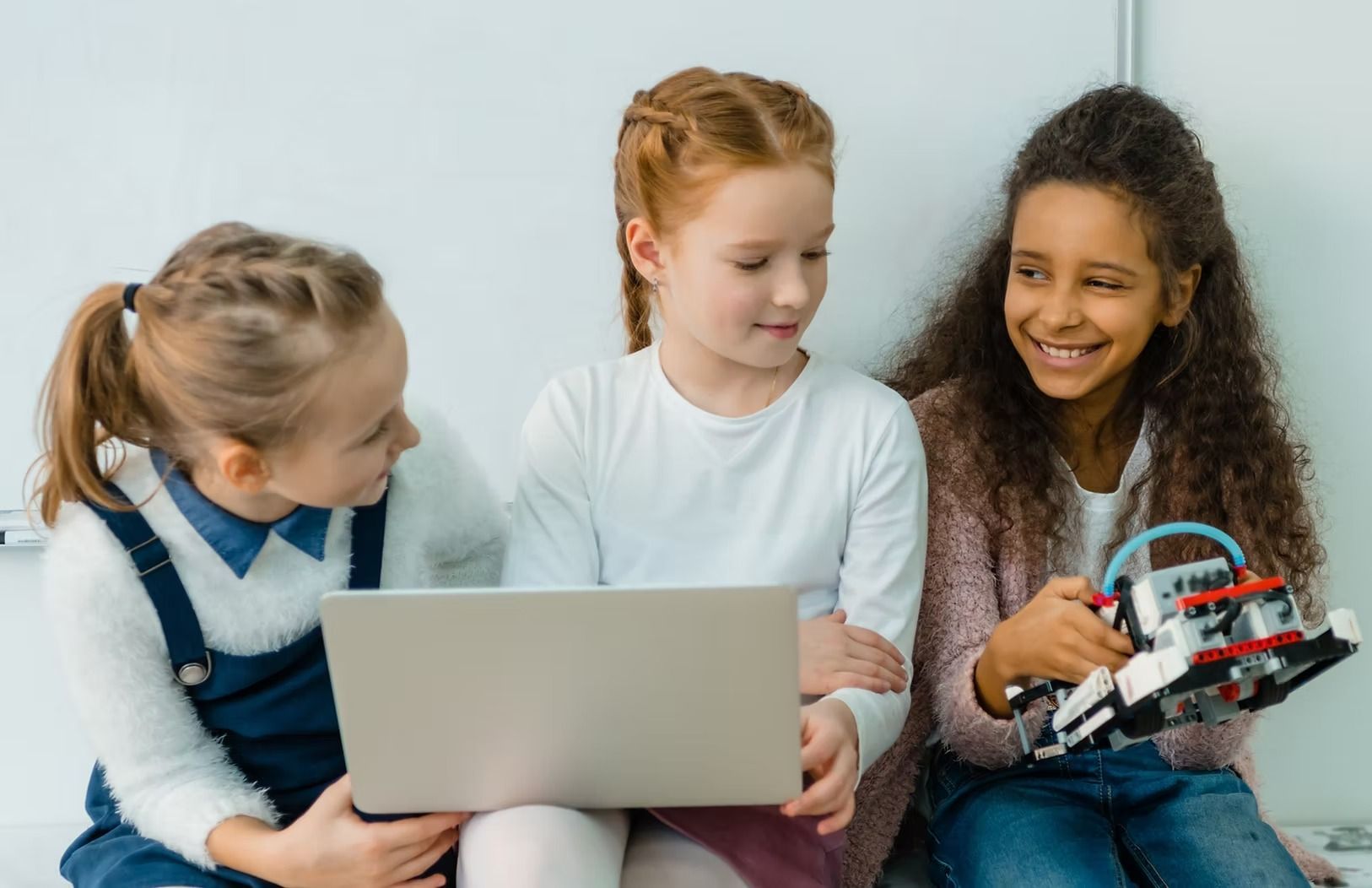 Three girls collaborate, looking at a laptop and robot. One girl holds the robot, smiling. Bright room.