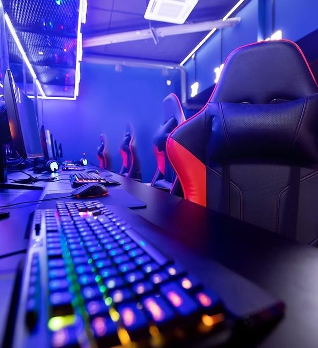Gaming chairs and keyboards in a dimly lit room with blue and purple LED lights.