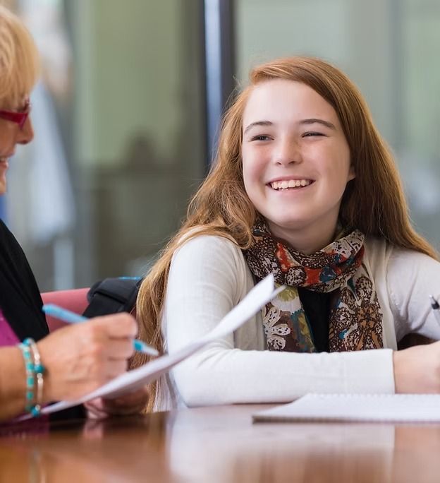 Teenager smiles while seated at a table, looking at papers with a woman. Both appear to be in a classroom.