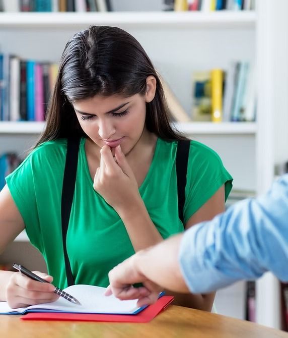Woman in green top studying at desk, looking at paper. Someone pointing. Bookshelf background.
