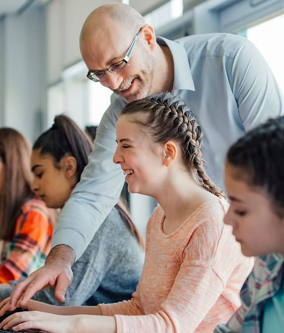 Teacher assisting a student at a computer, smiling. Other students seated nearby in a classroom.