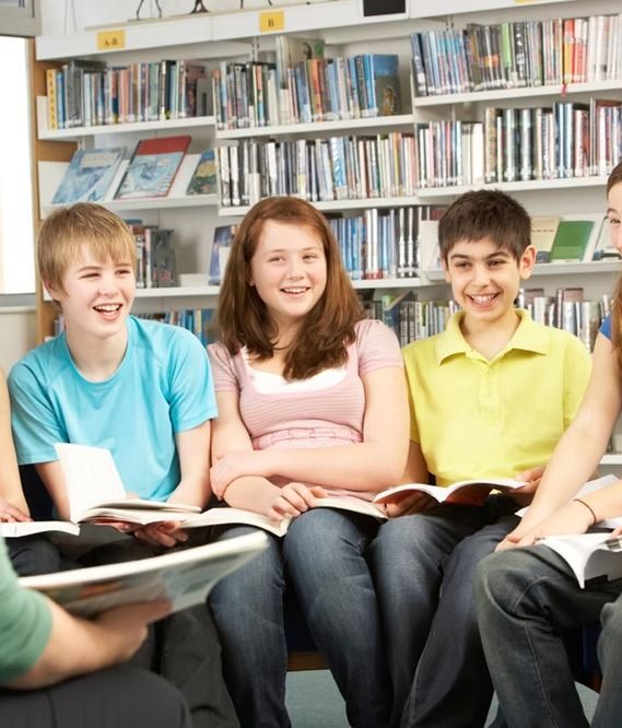 Group of teens smiling, reading books in a library, bookshelves in background.