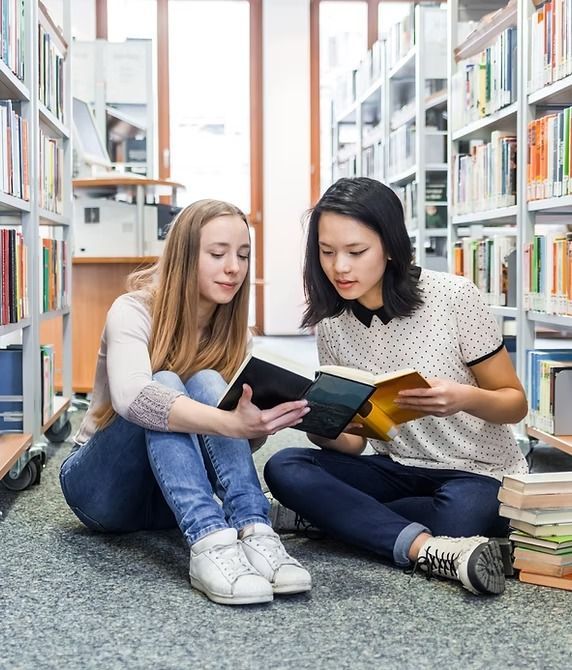 Two people sit on the library floor, reading books together. Bookshelves surround them.