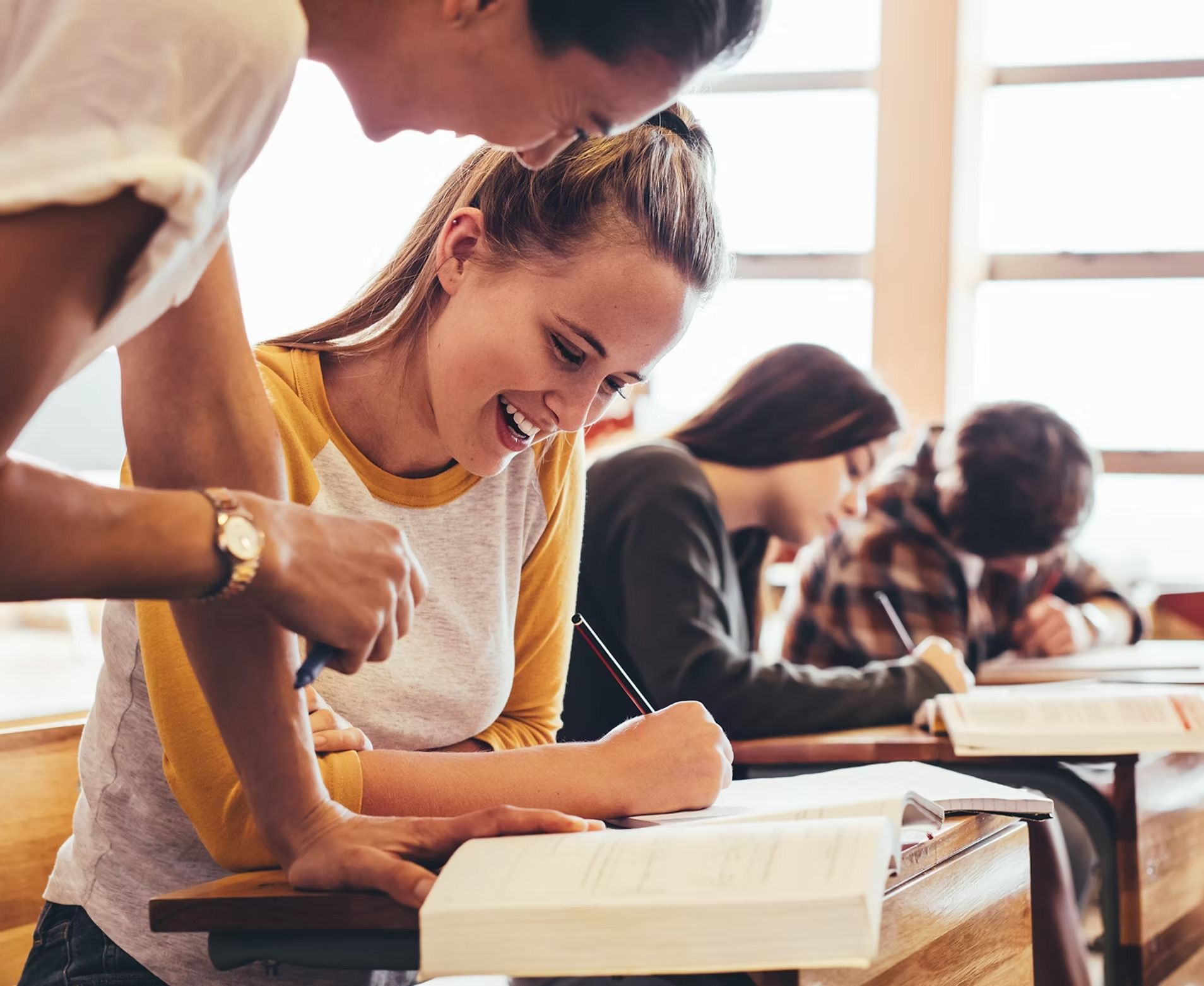 Two students collaborating at a desk in a classroom, one smiling while writing. Others work at desks in background.