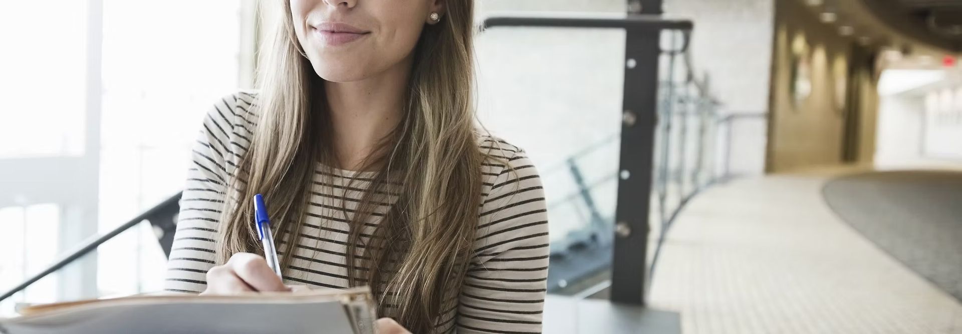 Woman writing on a notepad with a pen; a blurred hallway is in the background.