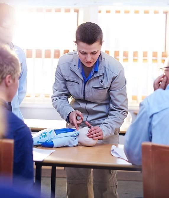 Person demonstrating on a white training dummy, with other people seated at a table, indoors.