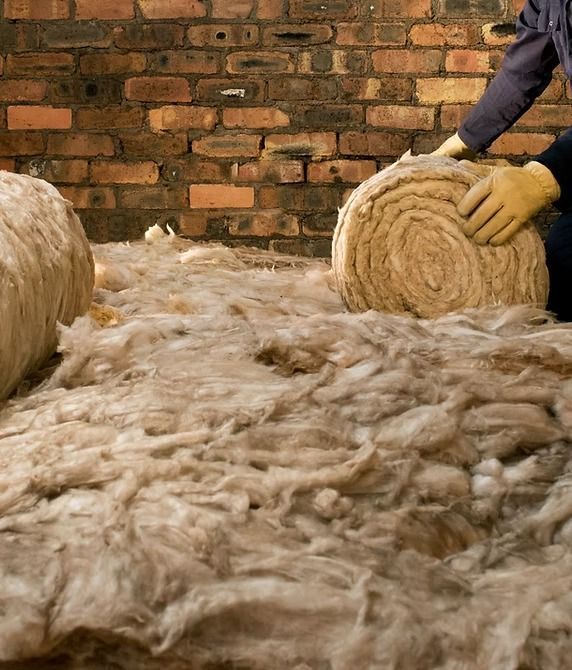 Person installing insulation rolls in a brick-walled room. Beige insulation is spread on the floor.