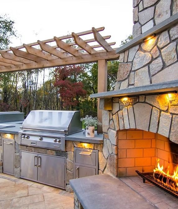 Outdoor kitchen with stainless steel grill, stone fireplace, and wooden pergola.