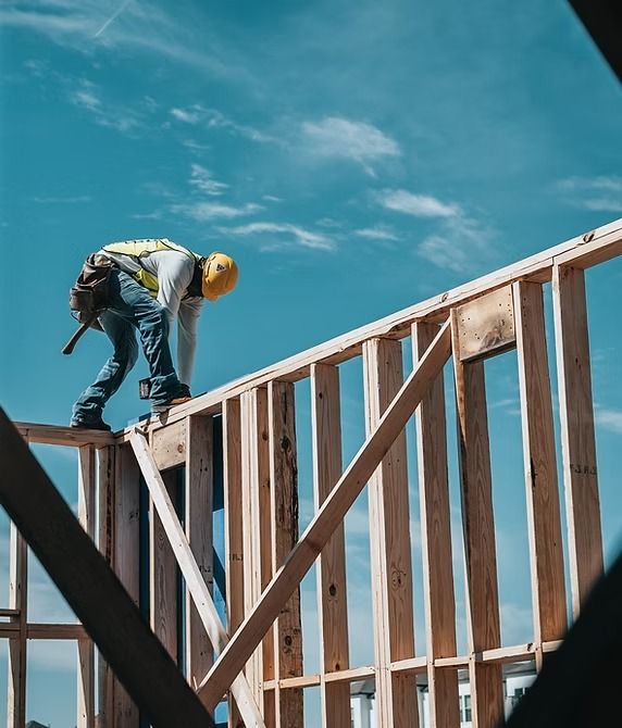 Construction worker on a wooden frame, wearing a yellow hard hat and safety vest, working under a blue sky.
