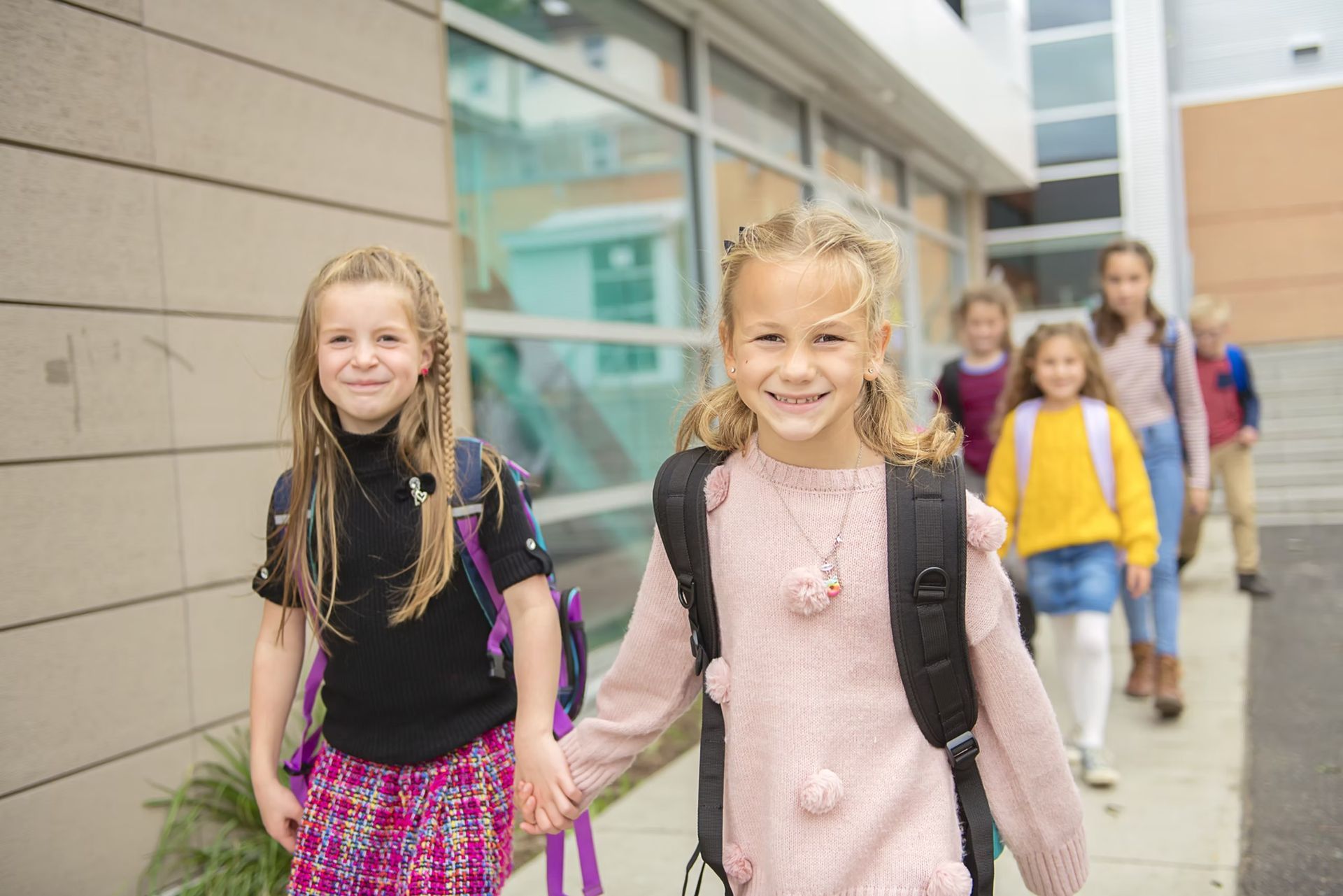 Children with backpacks walking outside a building, smiling.