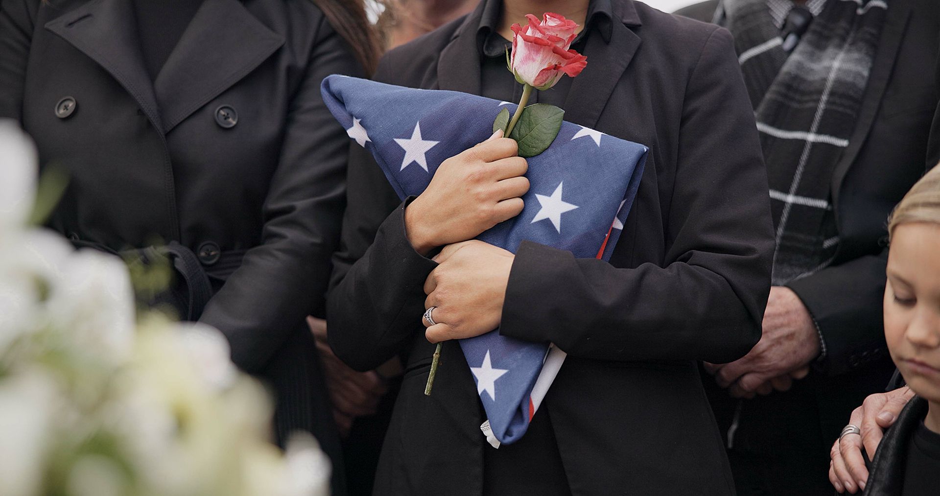 A person dressed in black holds a folded American flag and a rose during a somber funeral service.