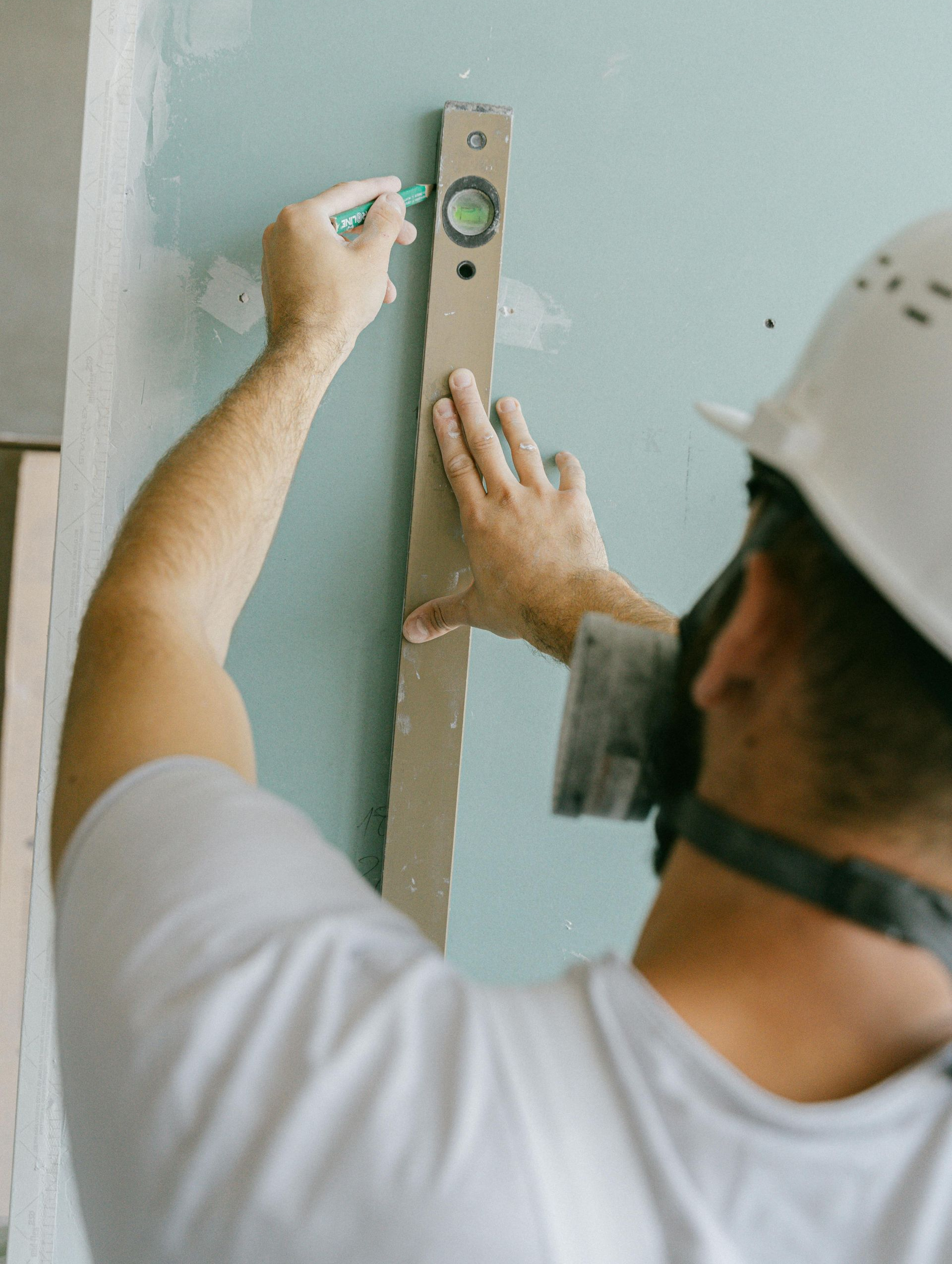 A worker in a hard hat and mask uses a spirit level and pencil to mark a light blue wall.