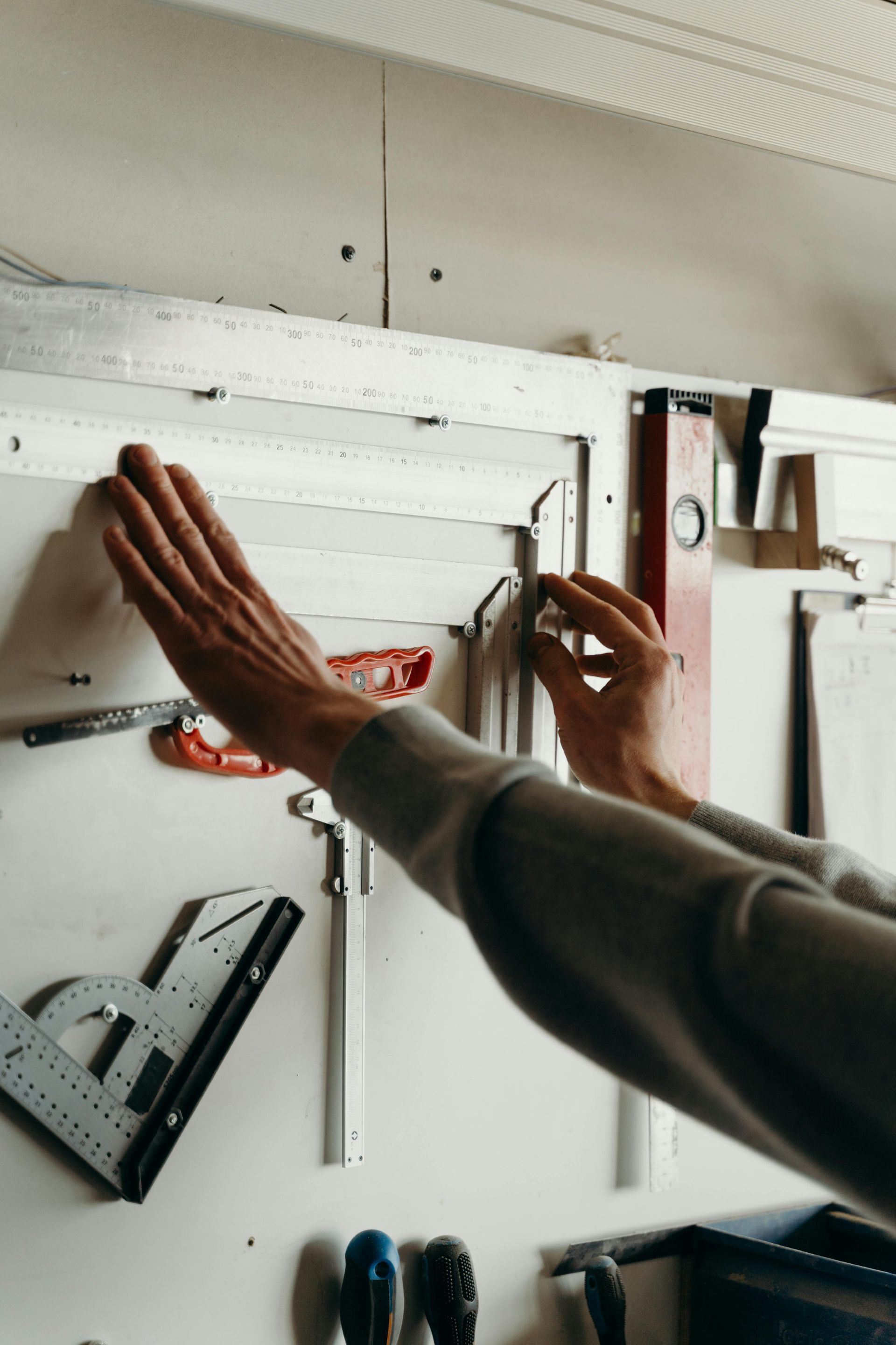 A person uses a metal ruler to measure a wooden frame attached to a white wall mounted with various workshop tools.