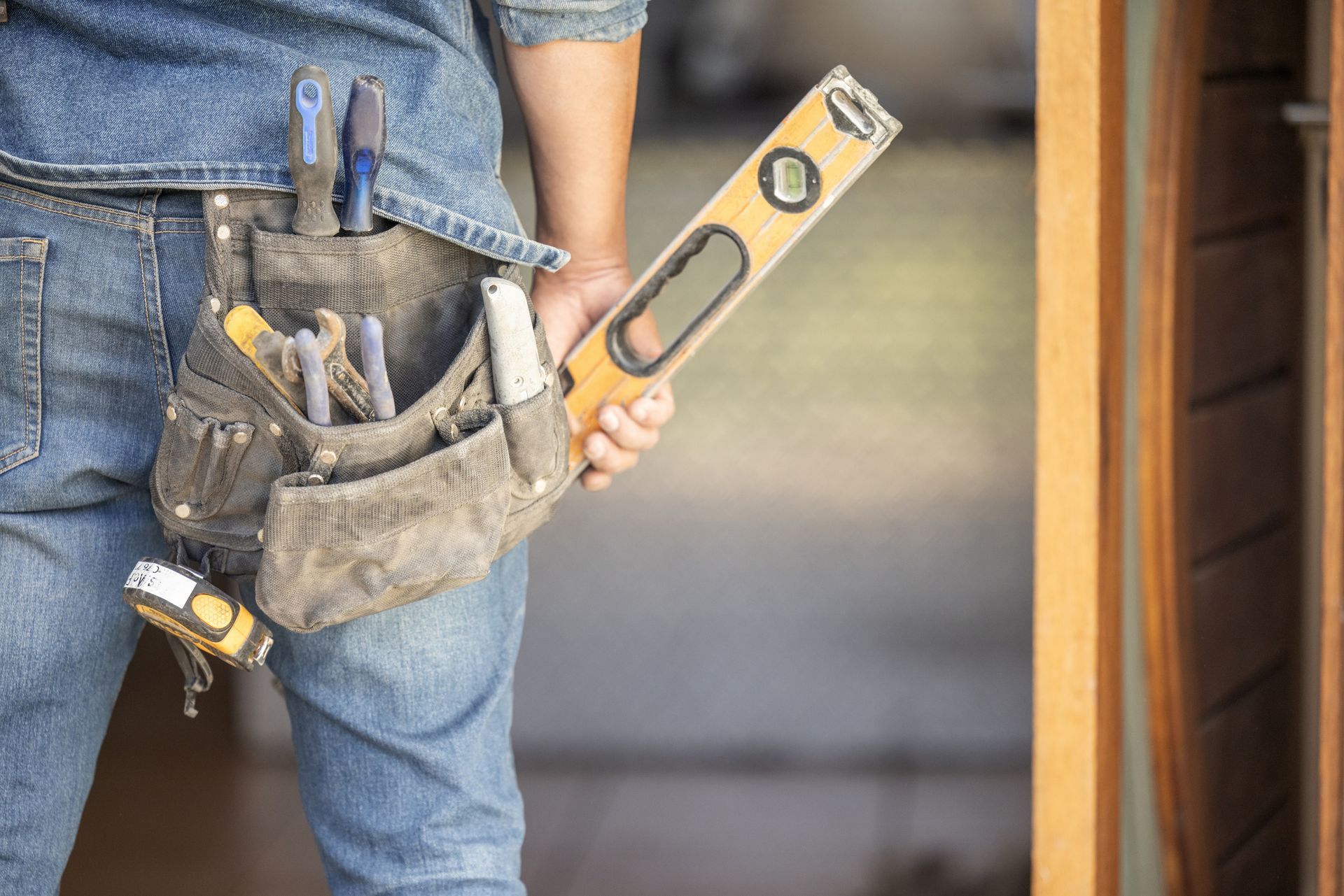 A person wearing a tool belt carries a carpenter's level while standing near a wooden door frame.