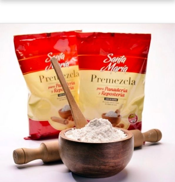 Dos bolsas de mezcla de harina Santa María, un cuenco de madera con harina y una cuchara, y un rodillo sobre un fondo blanco.