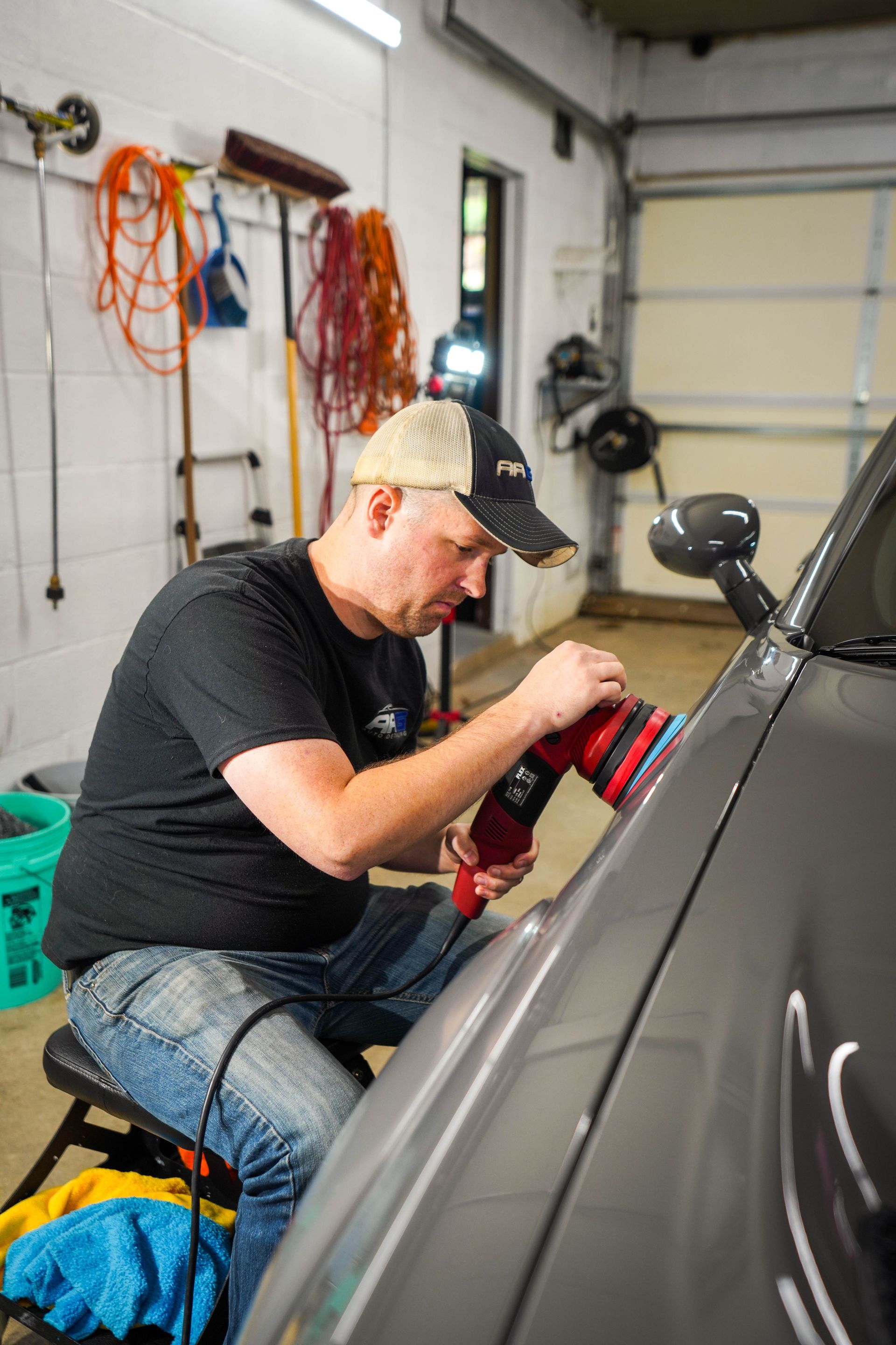Man polishes a gray car's side panel with a red power buffer in a garage.