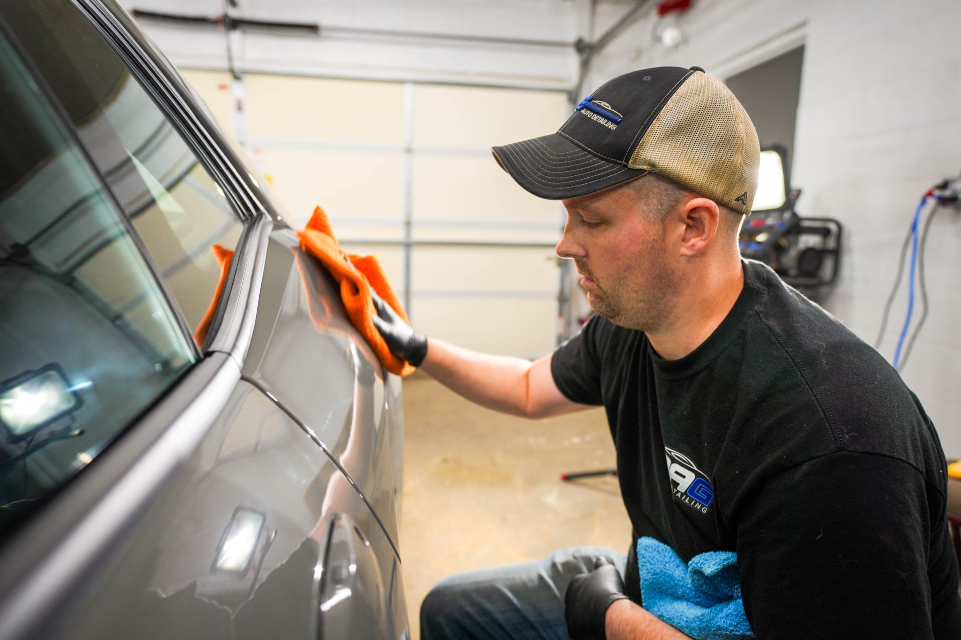 Man in hat and gloves, cleaning a car with an orange cloth in a garage.