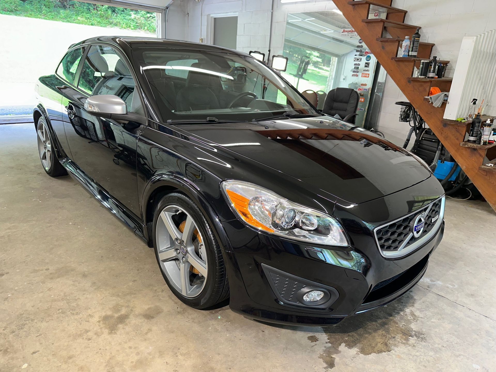 A black volvo c30 is parked in a garage next to a staircase.