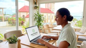 Une personne sourit en travaillant sur un ordinateur portable à une table dans une pièce ensoleillée