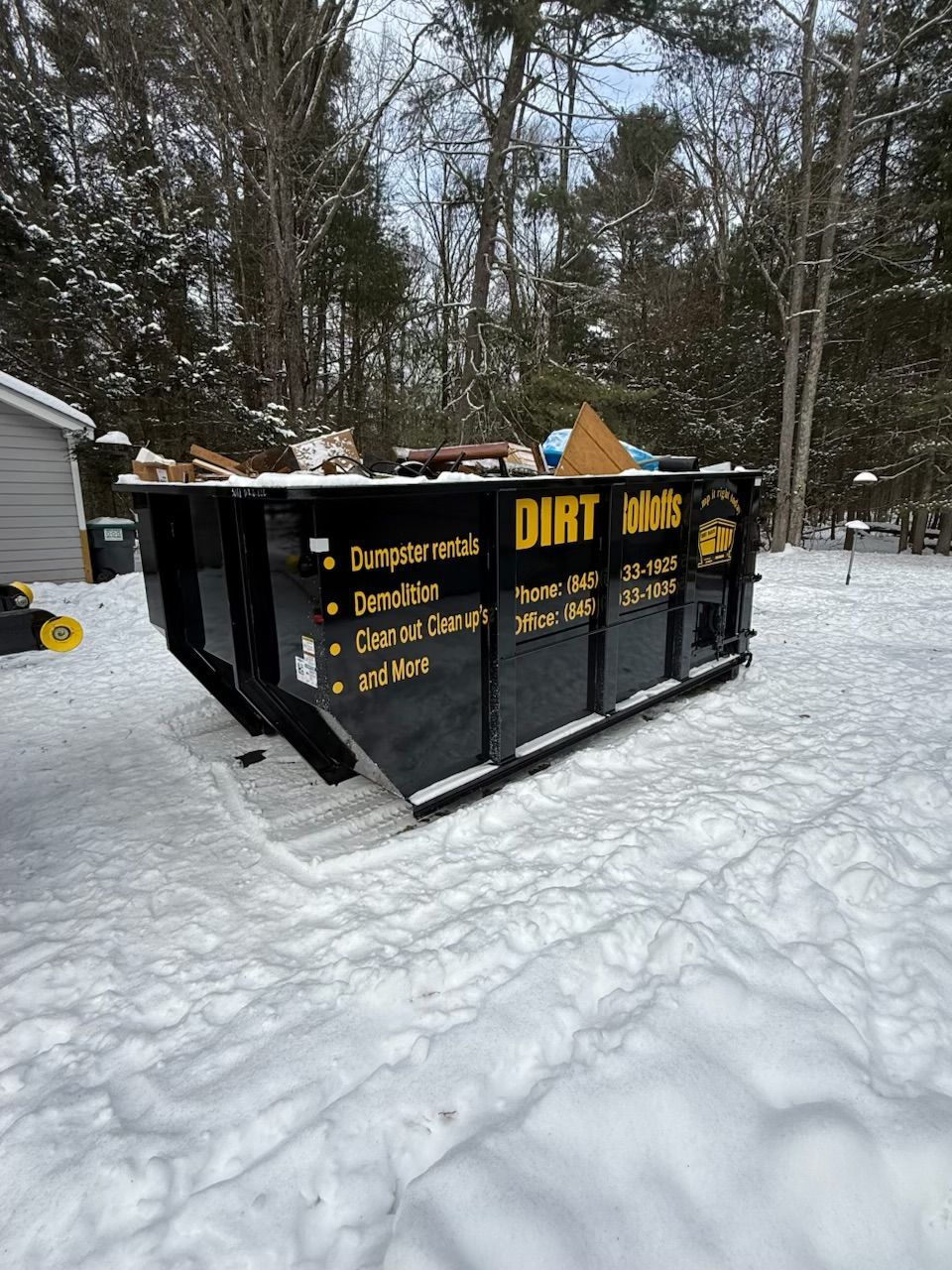 Black dumpster on snow-covered ground in front of a wooded area.
