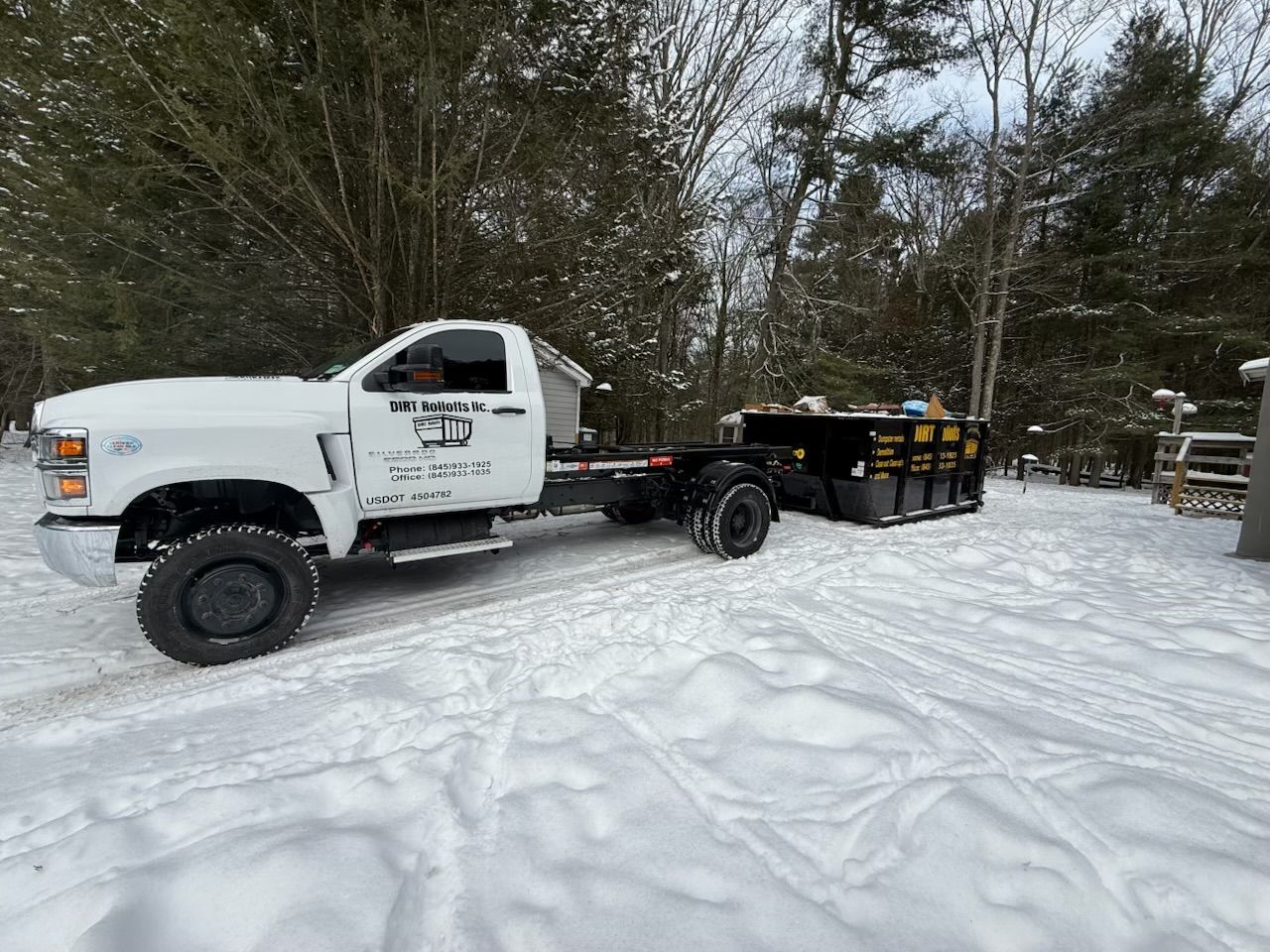 White truck pulling a trailer with black containers in a snowy, wooded area.