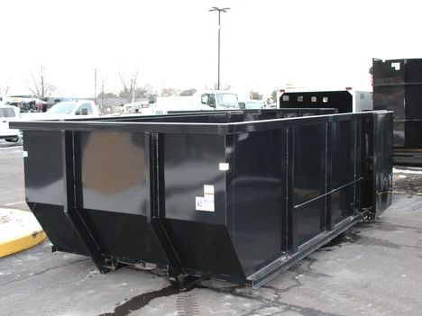 Black dumpster sits on green grass next to a house with a mailbox, trees, and wood.