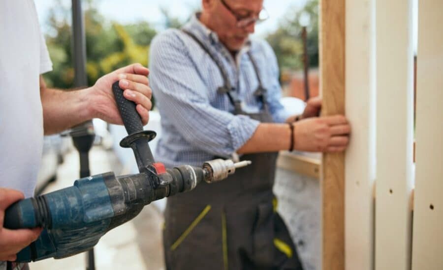 Two men building, one holding drill, one working with wood outdoors.