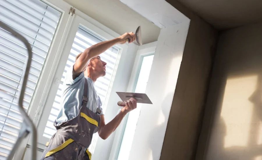 A man on a ladder is applying plaster to a wall with a trowel, near a window.