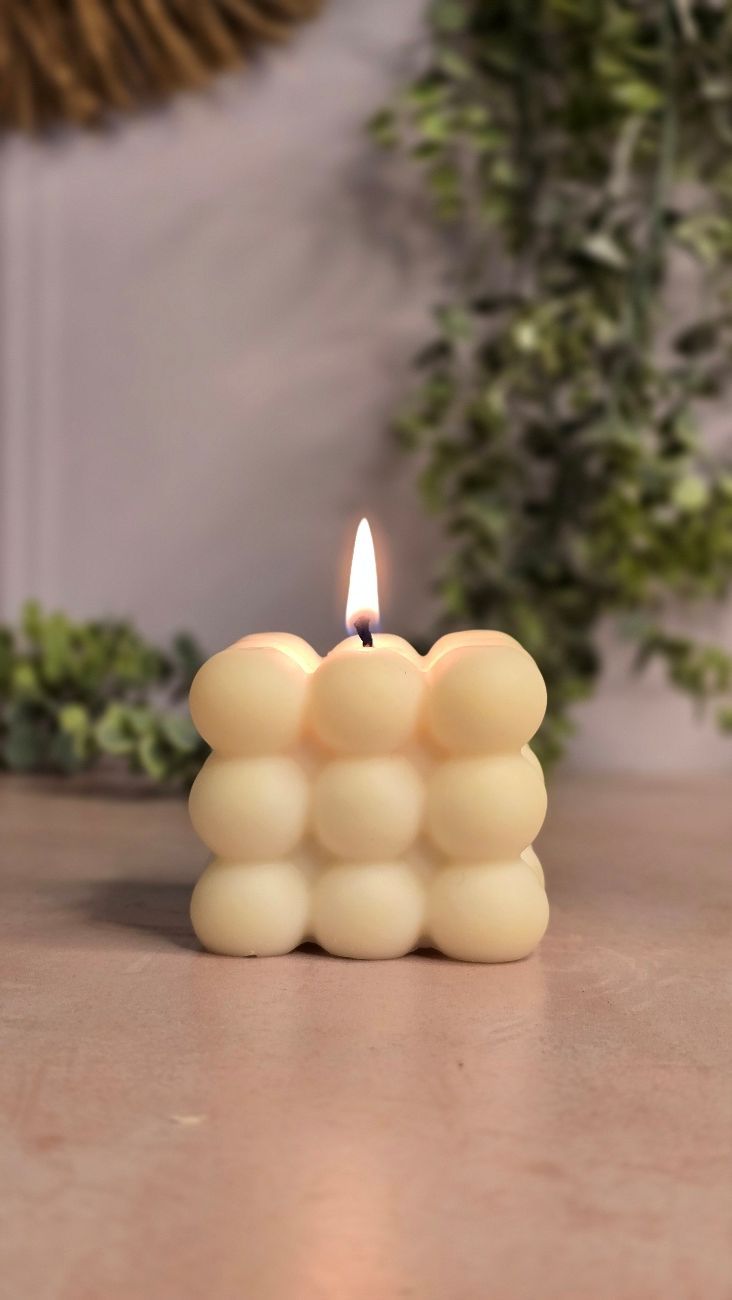 Vela de color crema encendida, con forma de burbuja, sobre una superficie de color claro con vegetación de fondo.