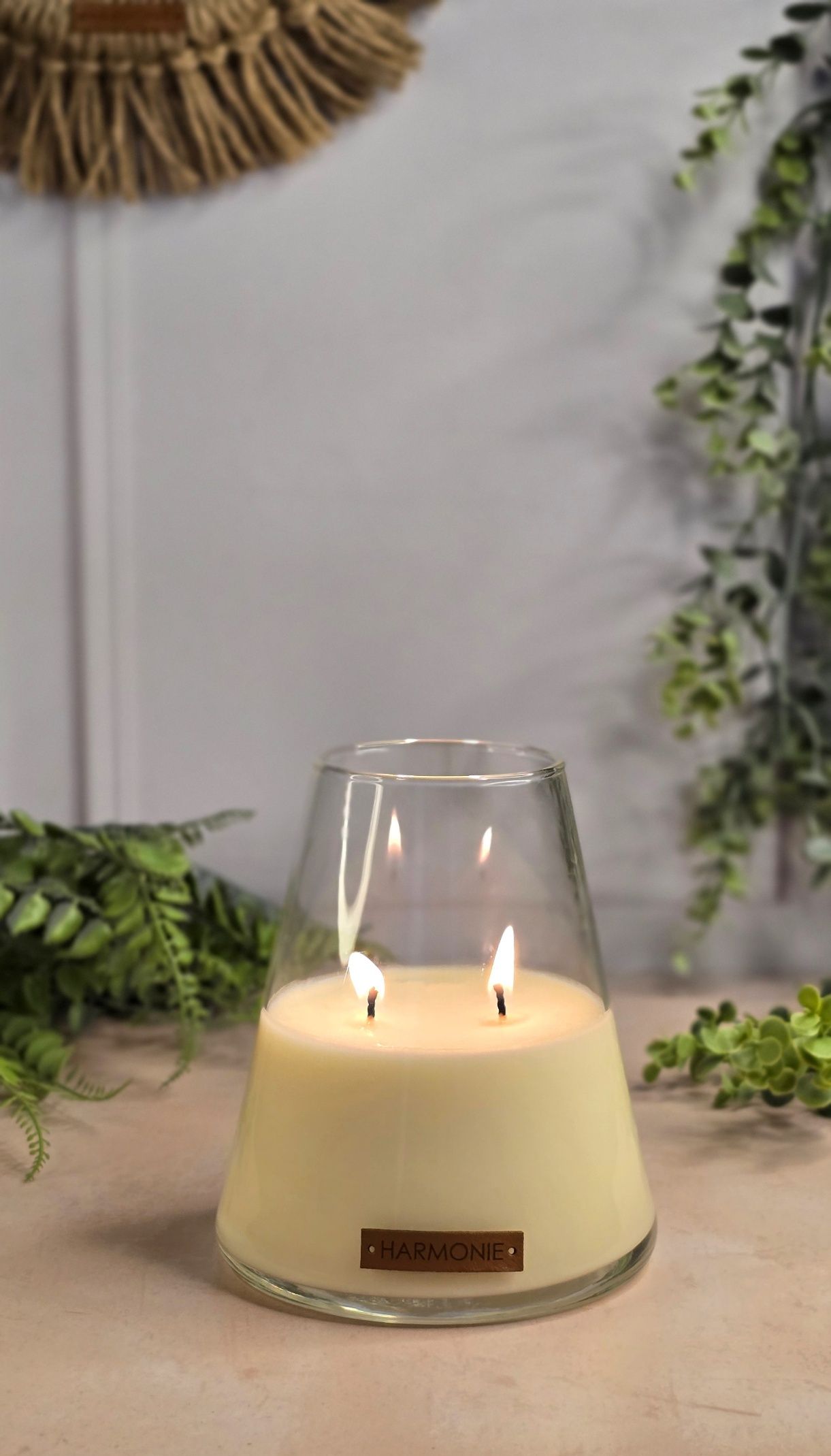 Una vela encendida de dos mechas en un recipiente de vidrio transparente, rodeada de plantas verdes, sobre una superficie clara.