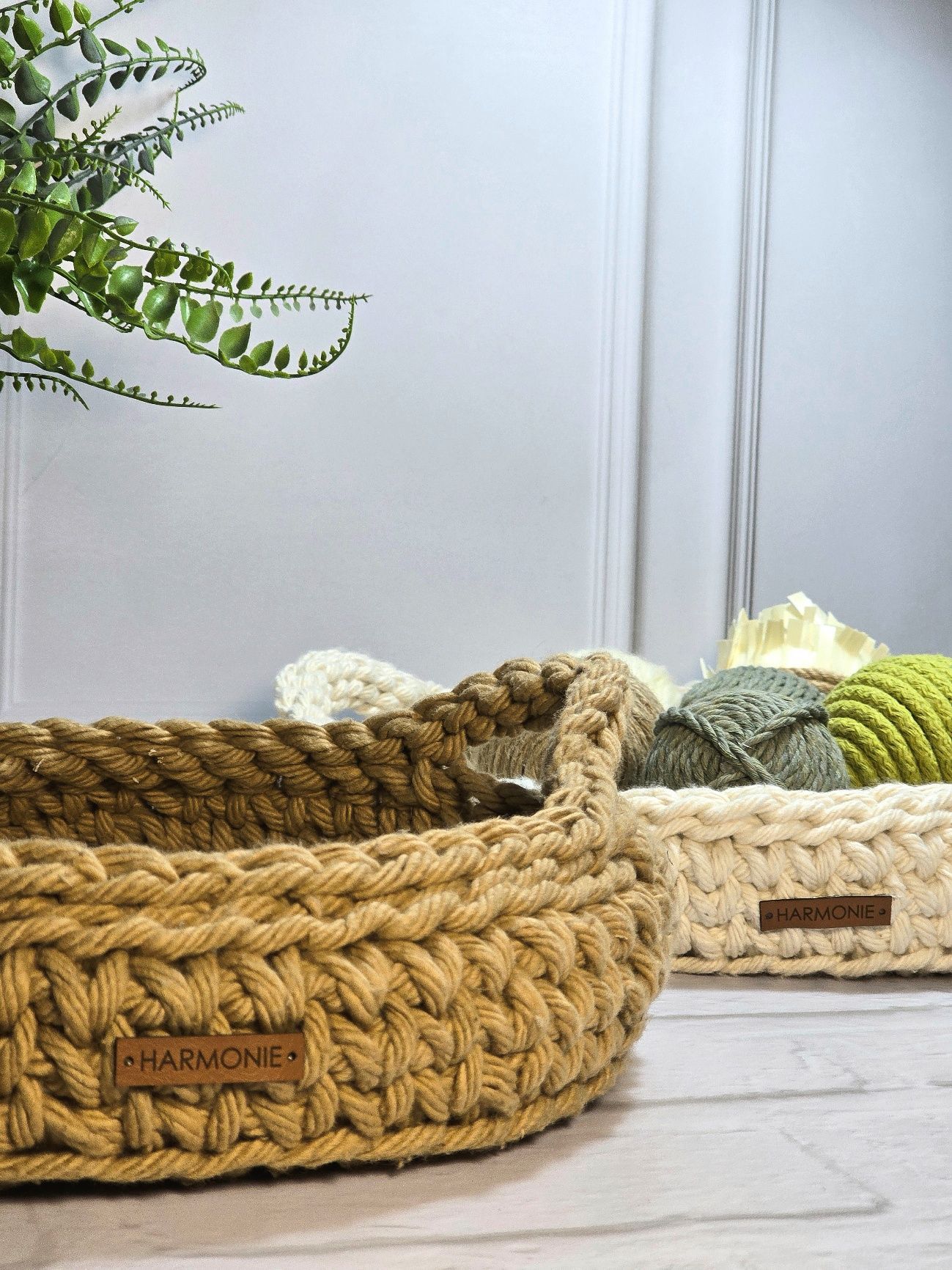 Dos cestas de ganchillo, de color beige y tostado, sobre una superficie de madera, con follaje verde de fondo.
