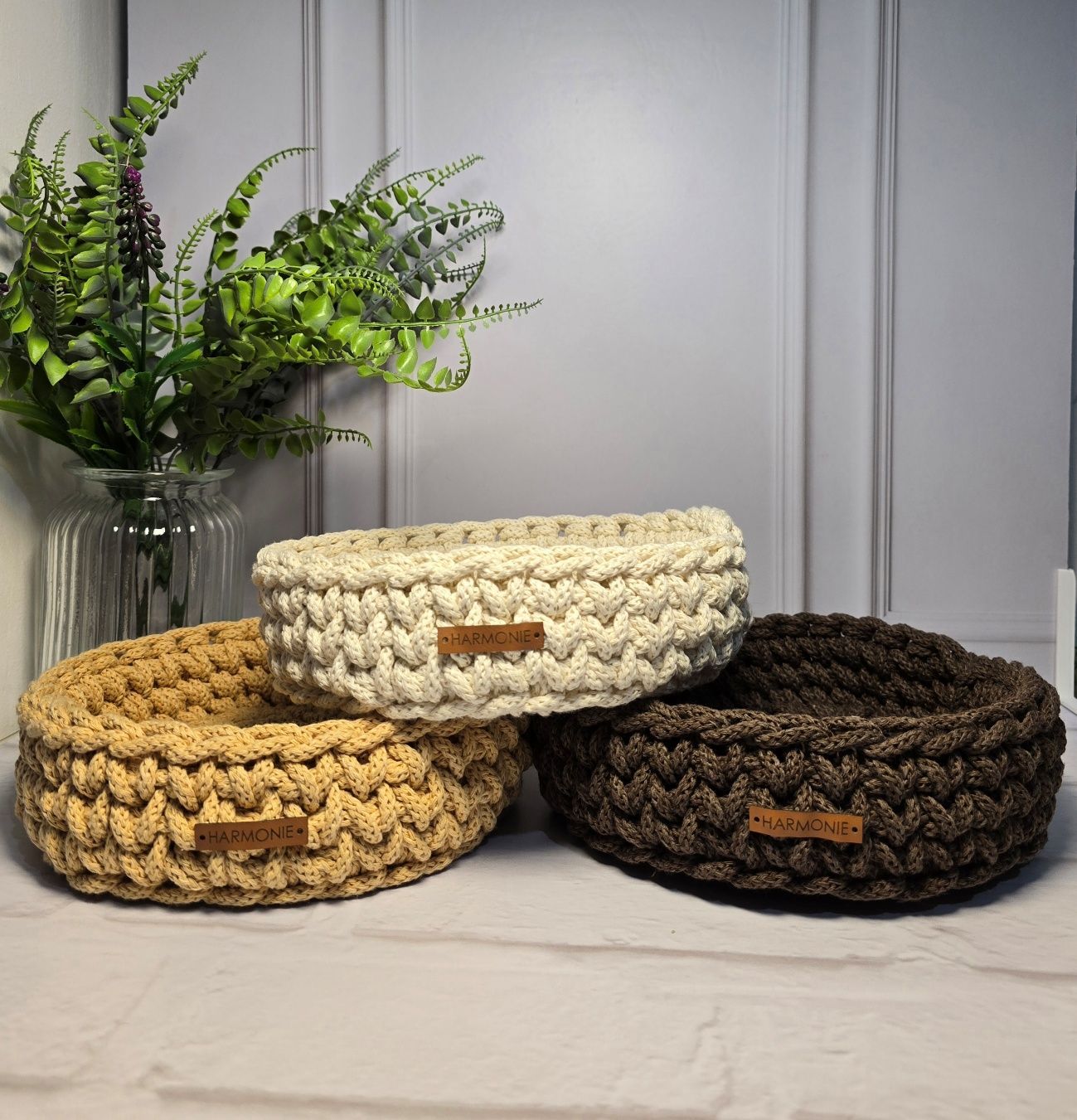 Tres cestas tejidas a crochet en tonos tostados, crema y marrón, con una planta en el fondo.