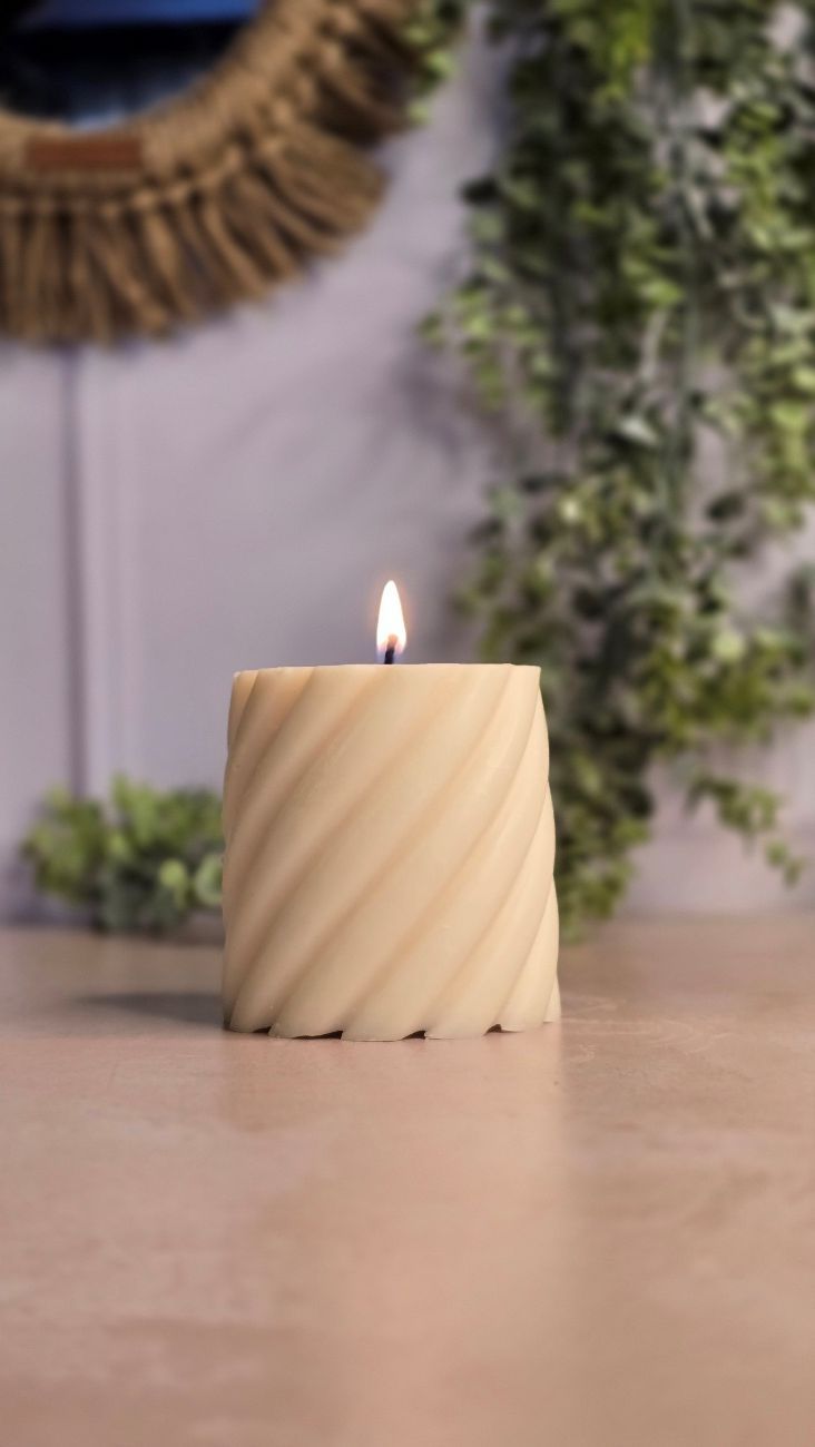 Una vela en espiral de color crema encendida sobre una mesa con vegetación y un espejo al fondo.