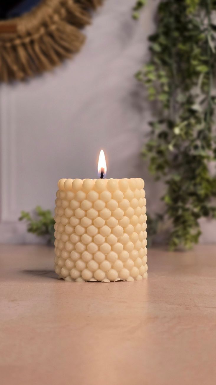 Vela encendida con textura de burbujas de color beige sobre una superficie de madera, con vegetación de fondo.