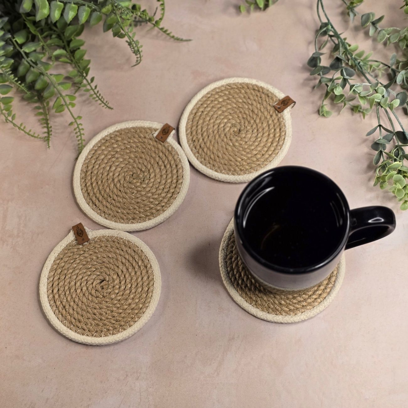 Cuatro posavasos redondos de yute con una taza negra, sobre una superficie beige rodeada de plantas.