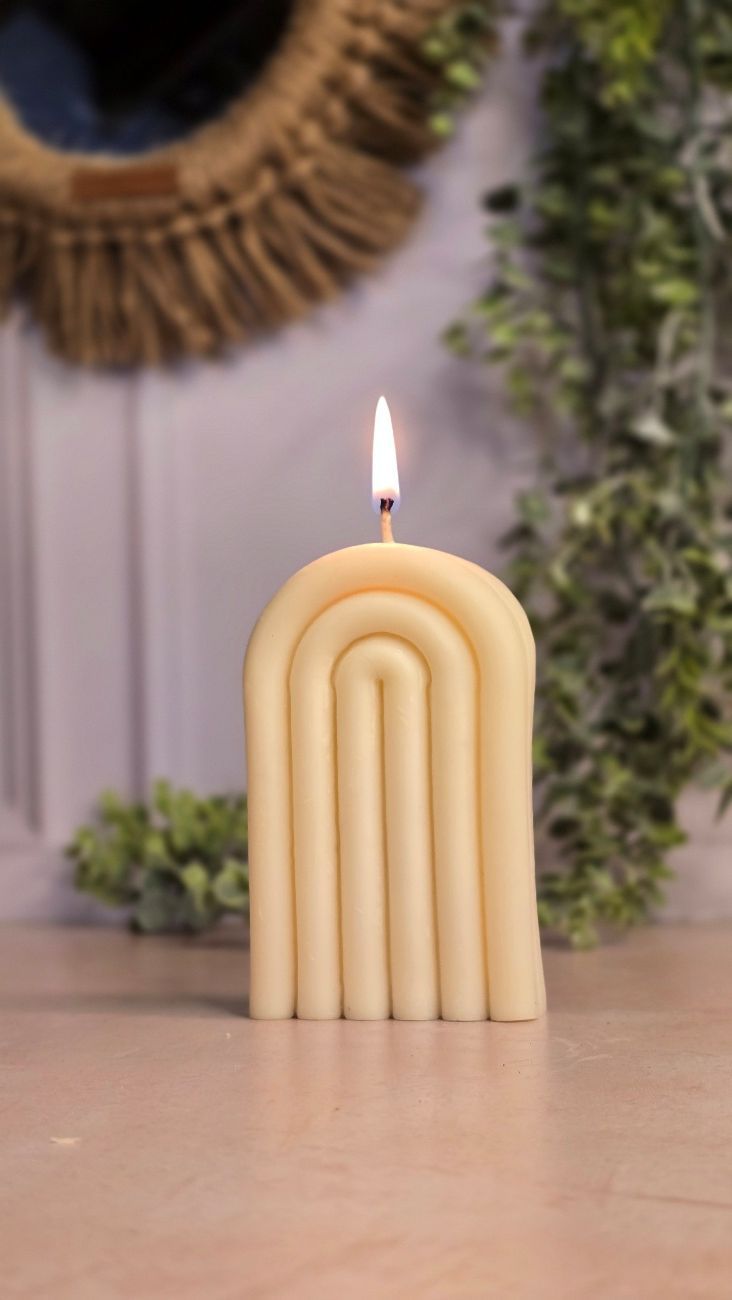 Vela encendida con forma de arcoíris sobre una superficie de madera con vegetación y un espejo tejido en el fondo.
