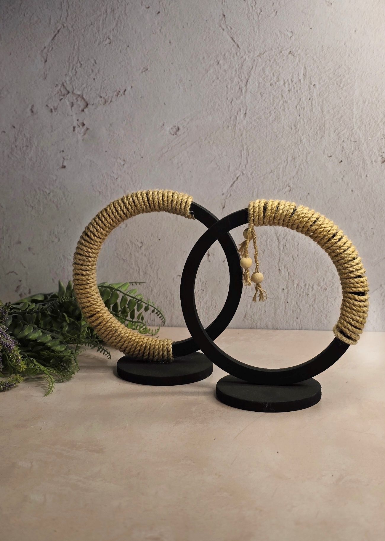 Dos piezas decorativas circulares negras, una de ellas envuelta en una cuerda color canela, sobre una superficie beige con follaje verde.