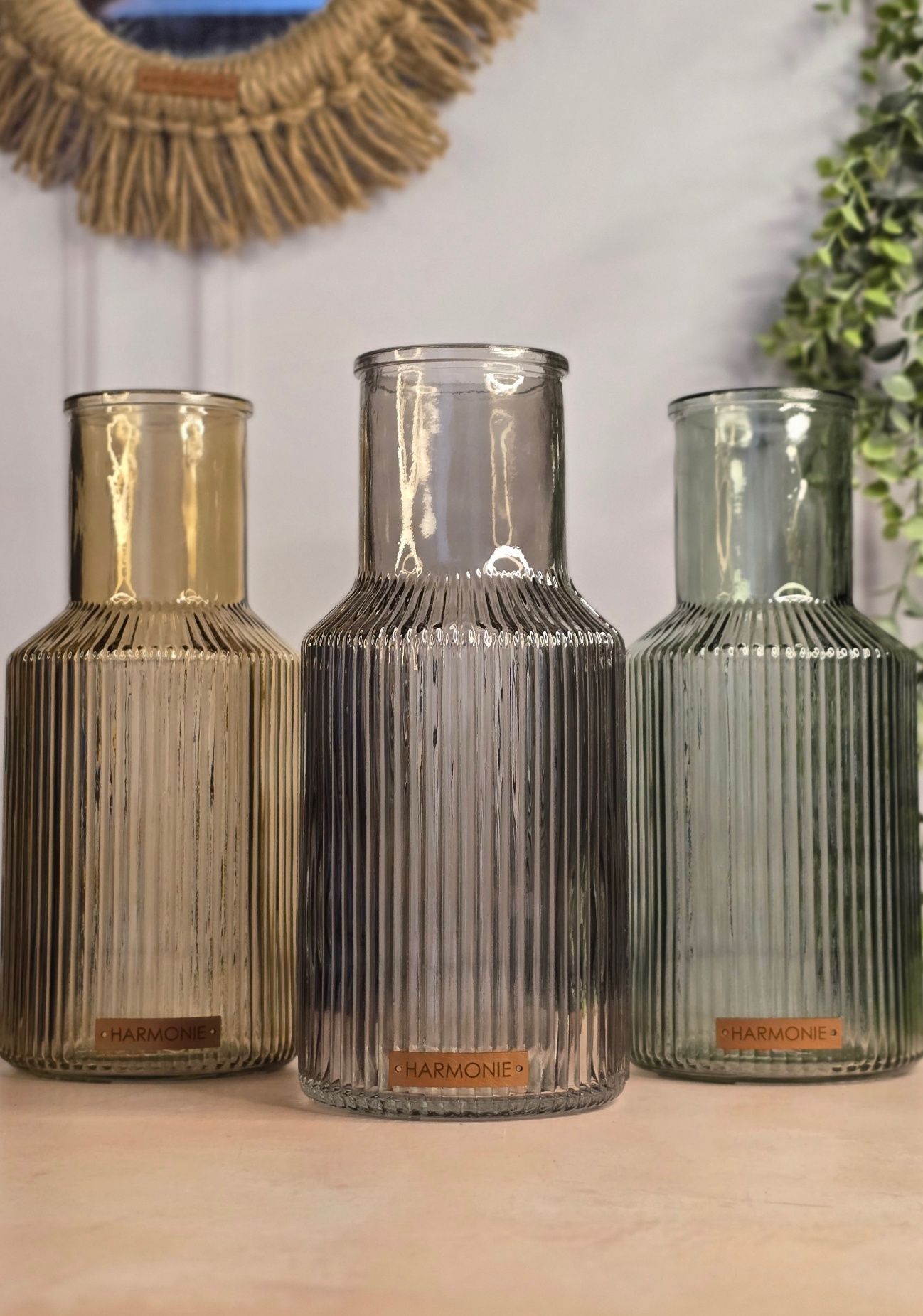 Tres jarrones de vidrio acanalado: ámbar, gris y verde, sobre una mesa, con un espejo de macramé al fondo.