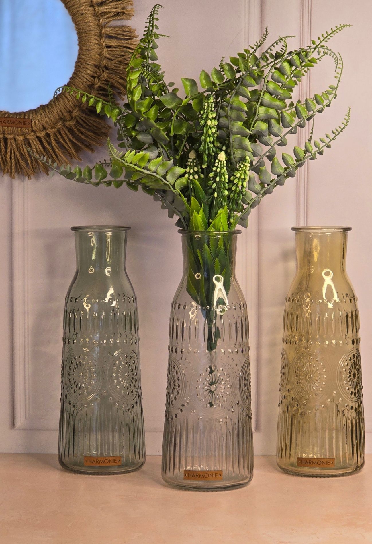 Tres jarrones de vidrio con follaje verde sobre una mesa frente a un espejo decorativo.