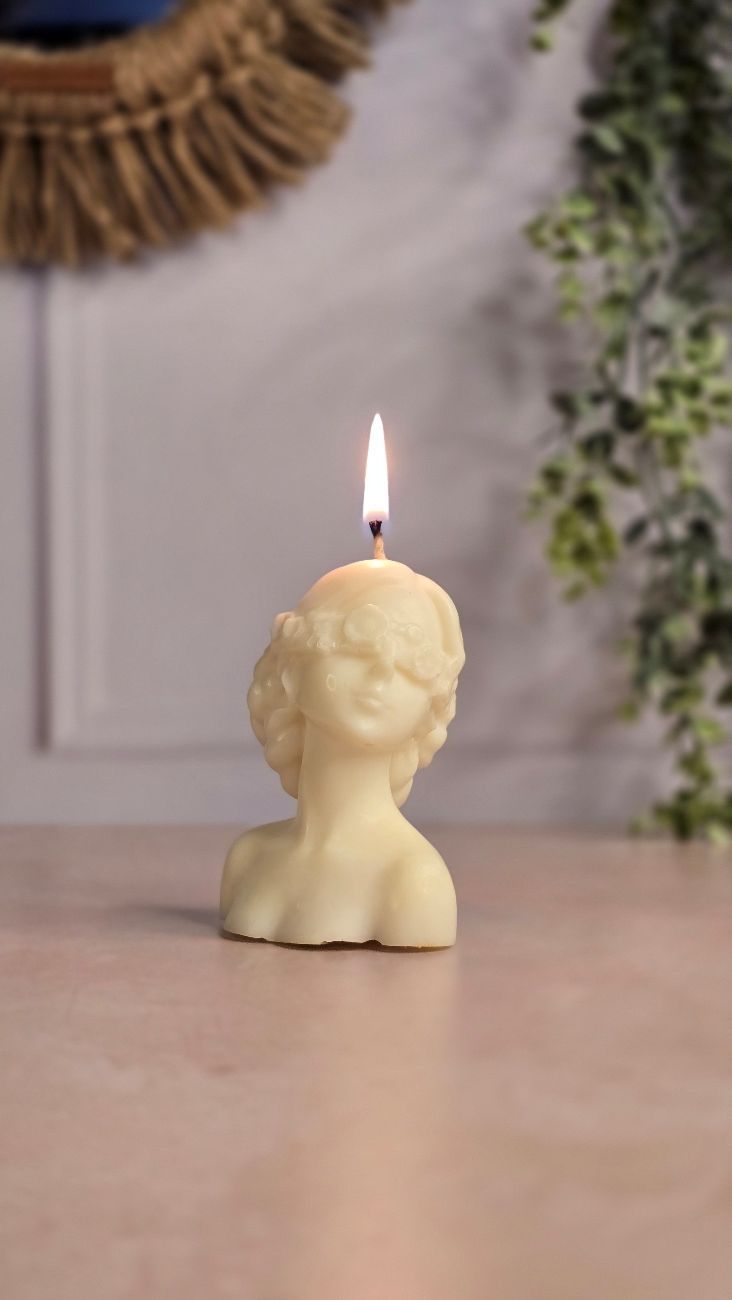 Vela ardiente de color crema con forma de busto de mujer sobre una superficie rosa; rodeada de vegetación.