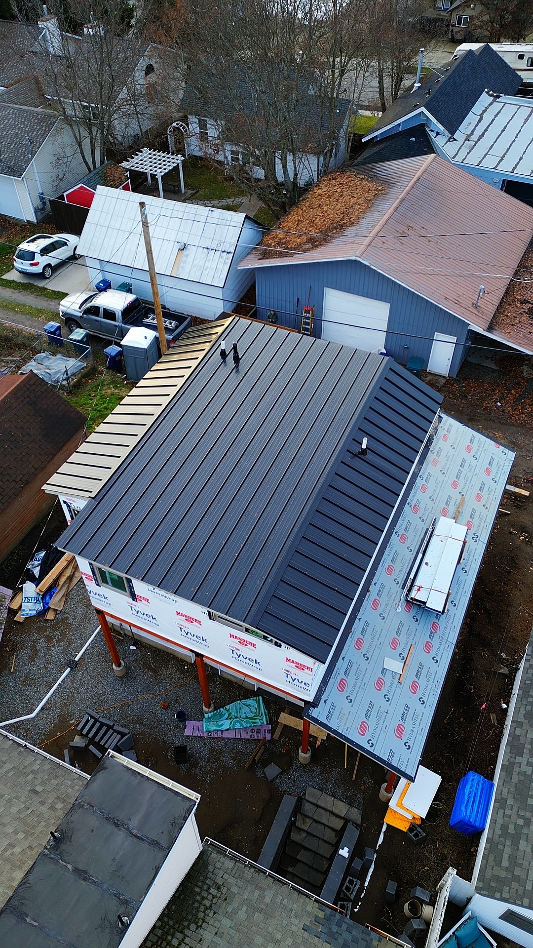 Aerial view of a house under construction with a black metal roof, surrounded by other buildings.